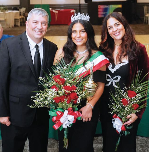 A person wearing a tiara and sash stands between two people, all smiling while holding bouquets in a formal setting.