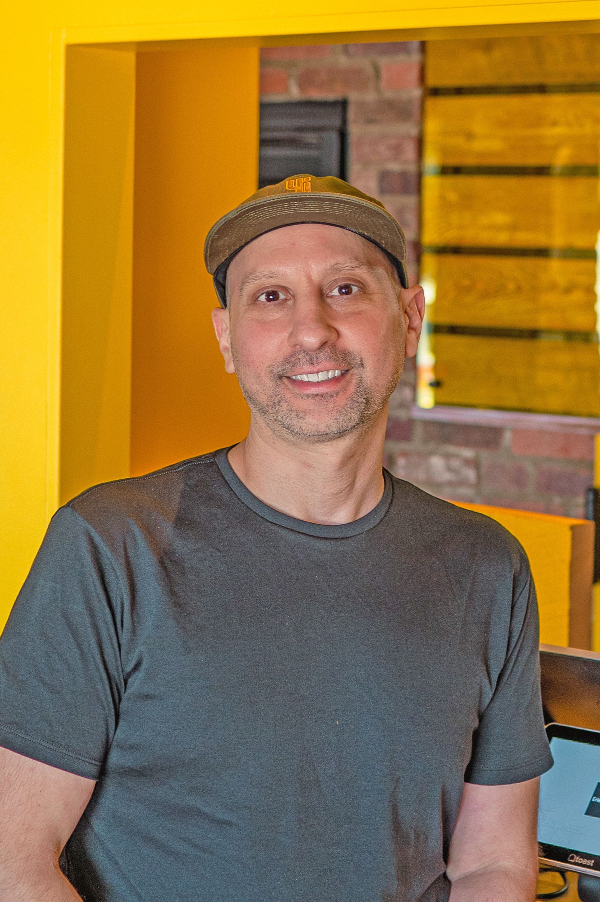 A smiling person wearing a grey t-shirt and a dark baseball cap, positioned in front of a yellow wall and brick background.