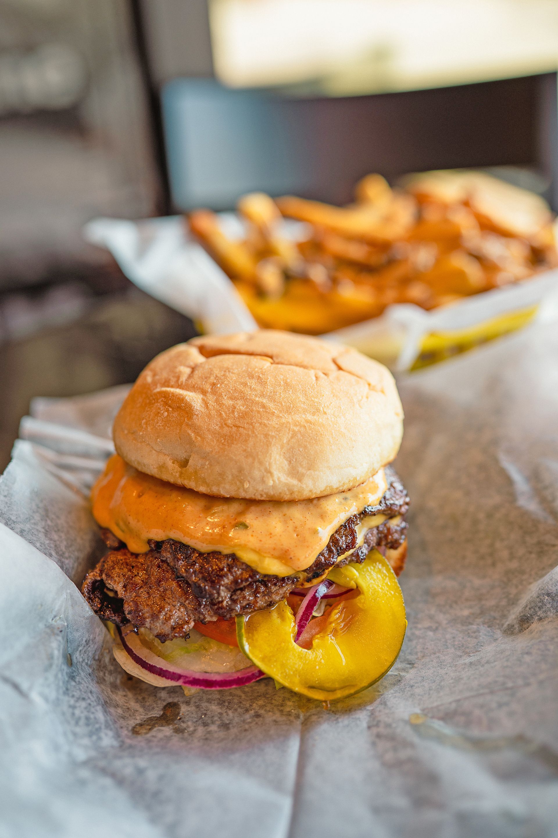 A close-up of a double cheeseburger with sauce, red onion, and pickles, served on paper with a side of fries.