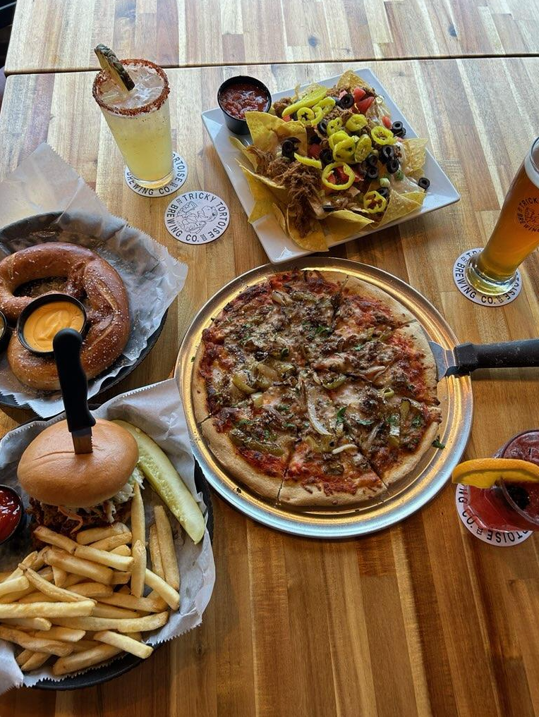 Top-down view of a wooden table set with a pepperoni pizza, nachos, a soft pretzel with dip, a burger with fries, and drinks.