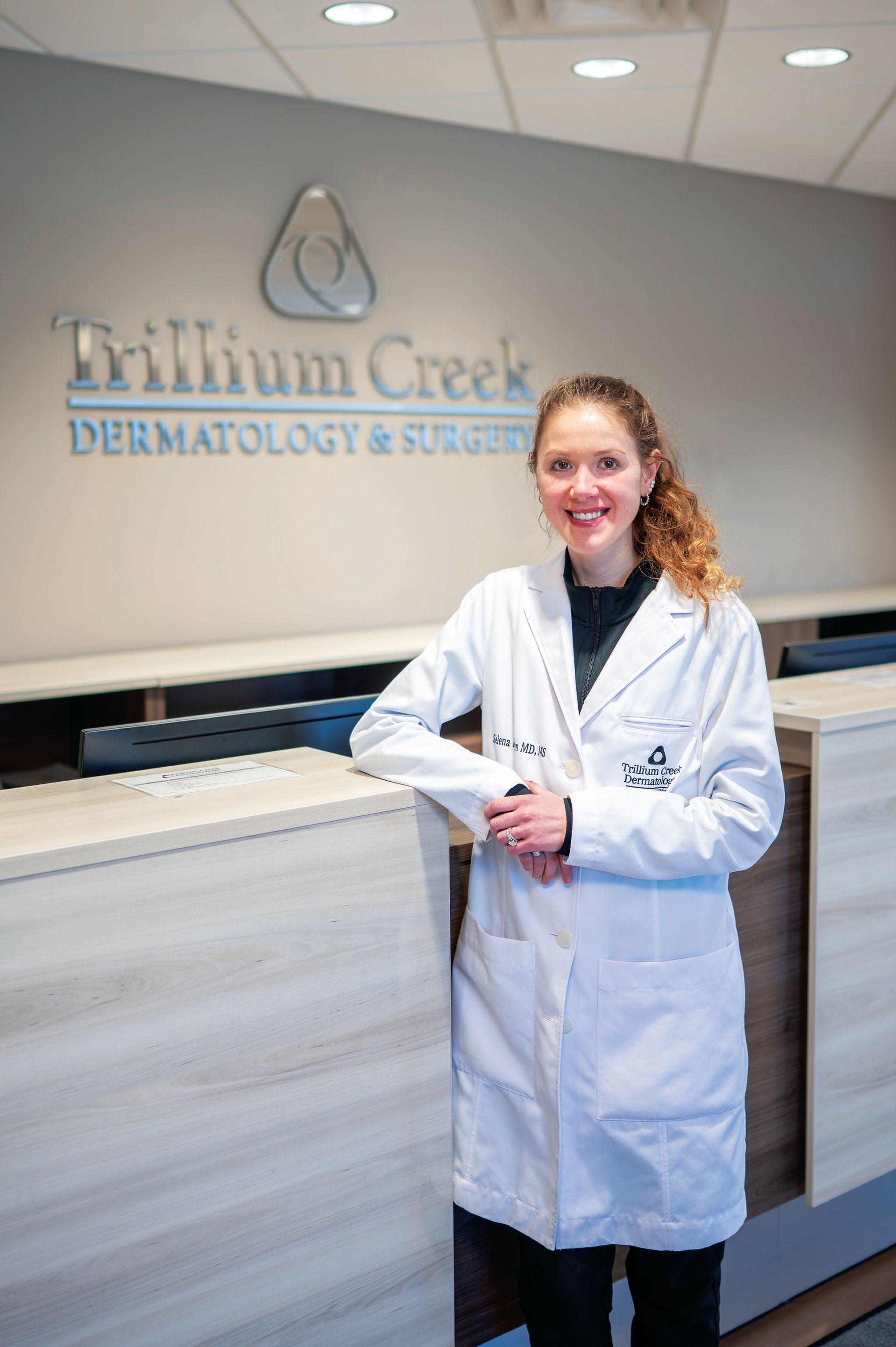 A person in a white medical coat smiles standing at the front desk of Trillium Creek Dermatology & Surgery.