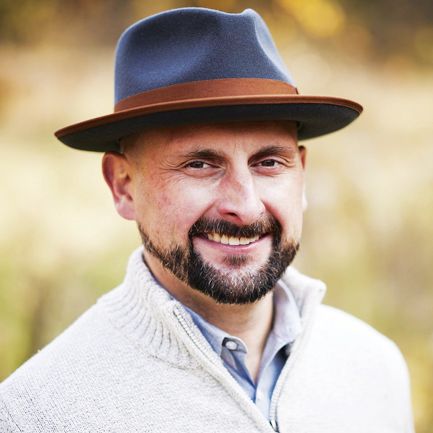 A person smiling, wearing a blue fedora with a brown band and a cream-colored knit sweater, set against a soft background.