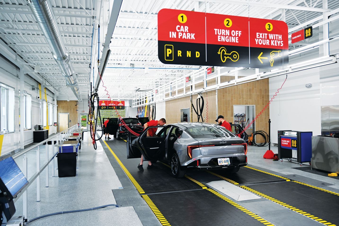 A silver car in a bright service bay with signage directing drivers to park, turn off the engine, and exit with keys.