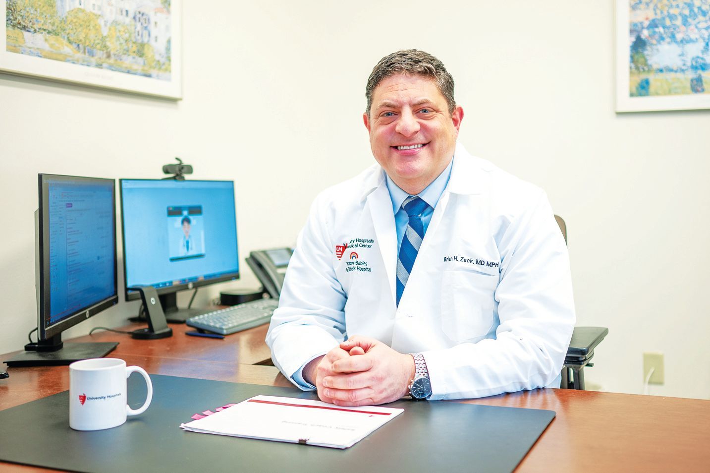 A smiling physician in a white coat sits at a desk with two computer monitors, a coffee mug, and papers.