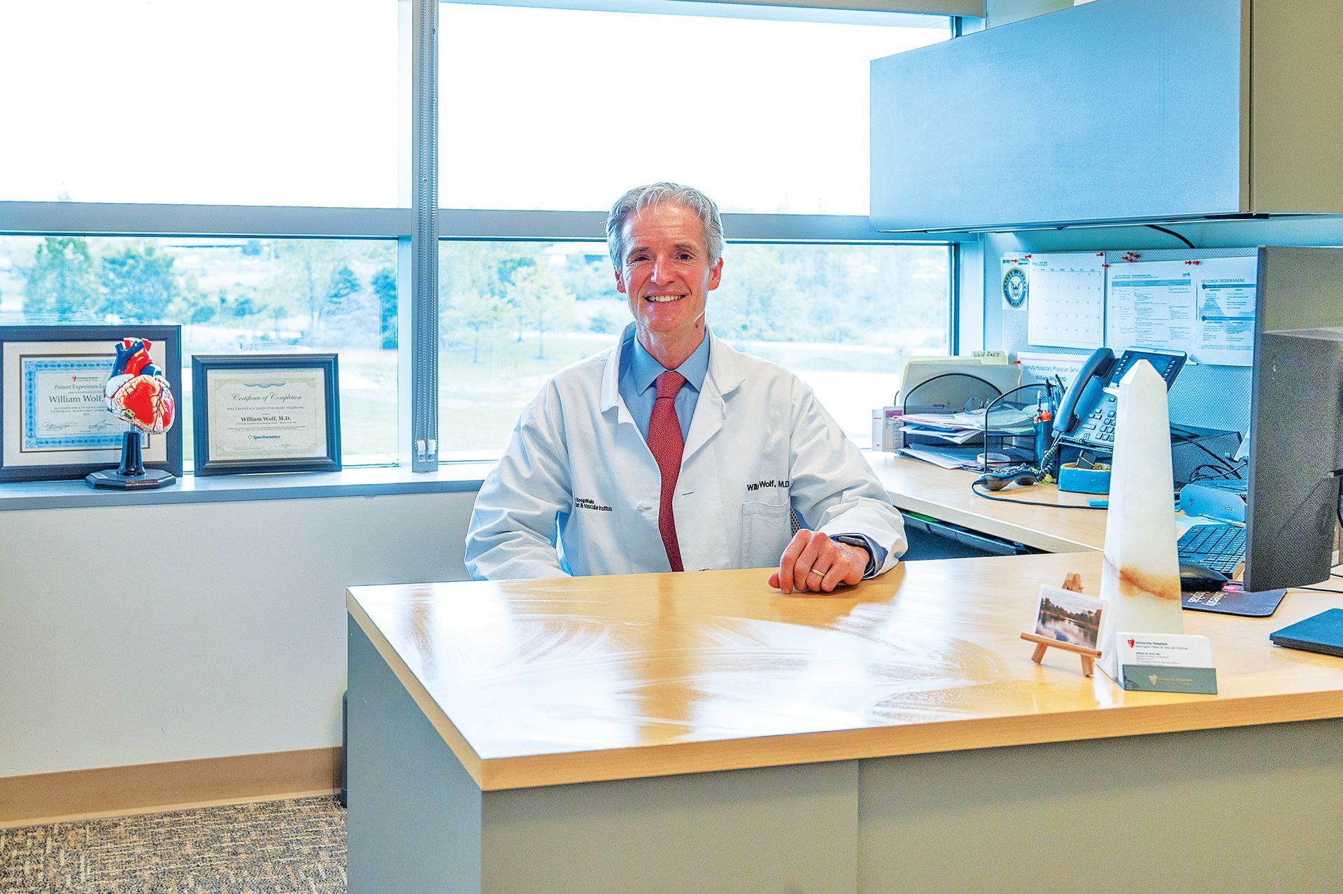 A person in a white medical coat smiles while sitting at a desk in an office with large windows.