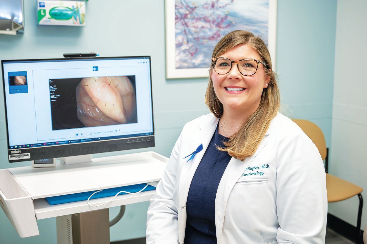 A smiling medical professional in a white coat stands next to a computer monitor displaying an endoscopic image.