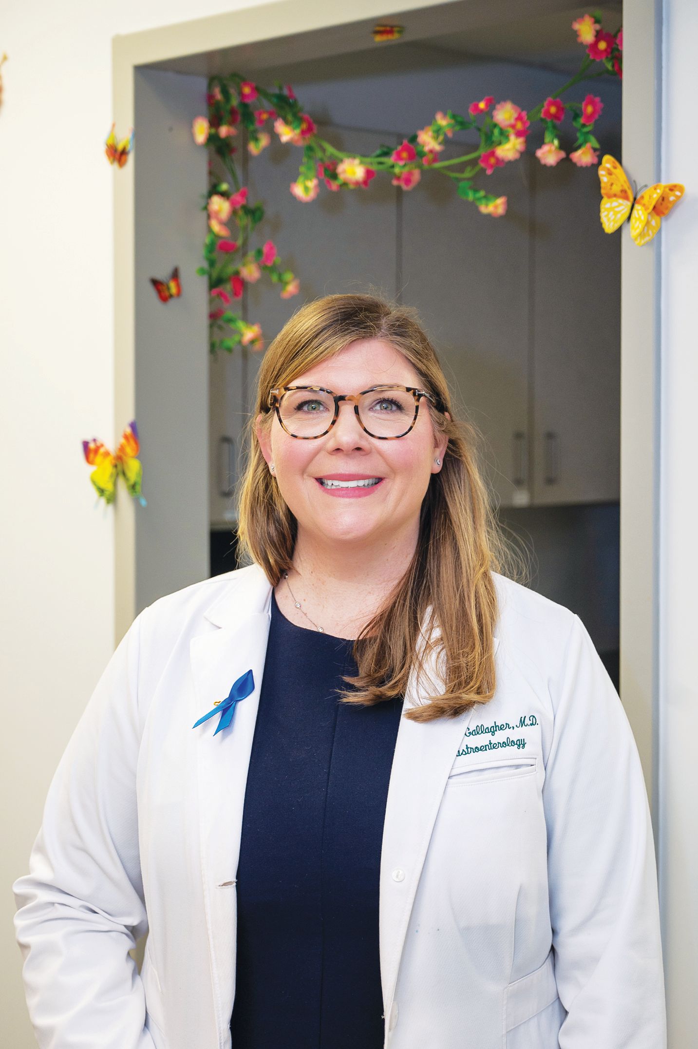 A professional in a white lab coat and glasses stands smiling in front of a doorway decorated with colorful butterflies.