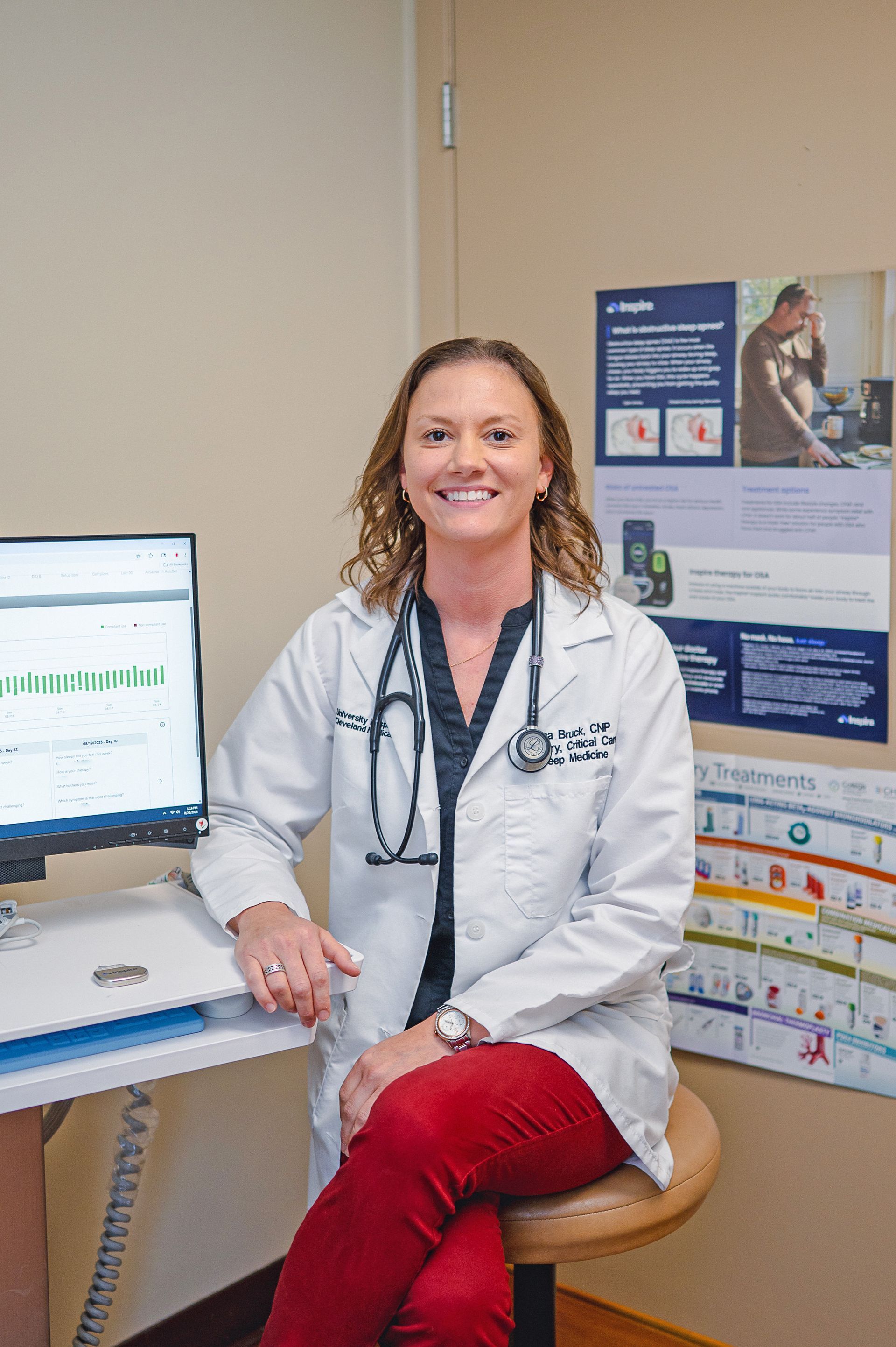 A professional in a white coat and stethoscope sits in a medical office next to a computer screen and wall charts.