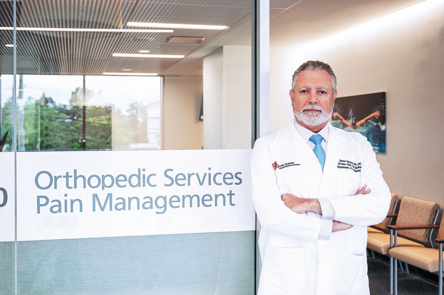 A physician in a white coat stands with arms crossed in front of a glass wall labeled