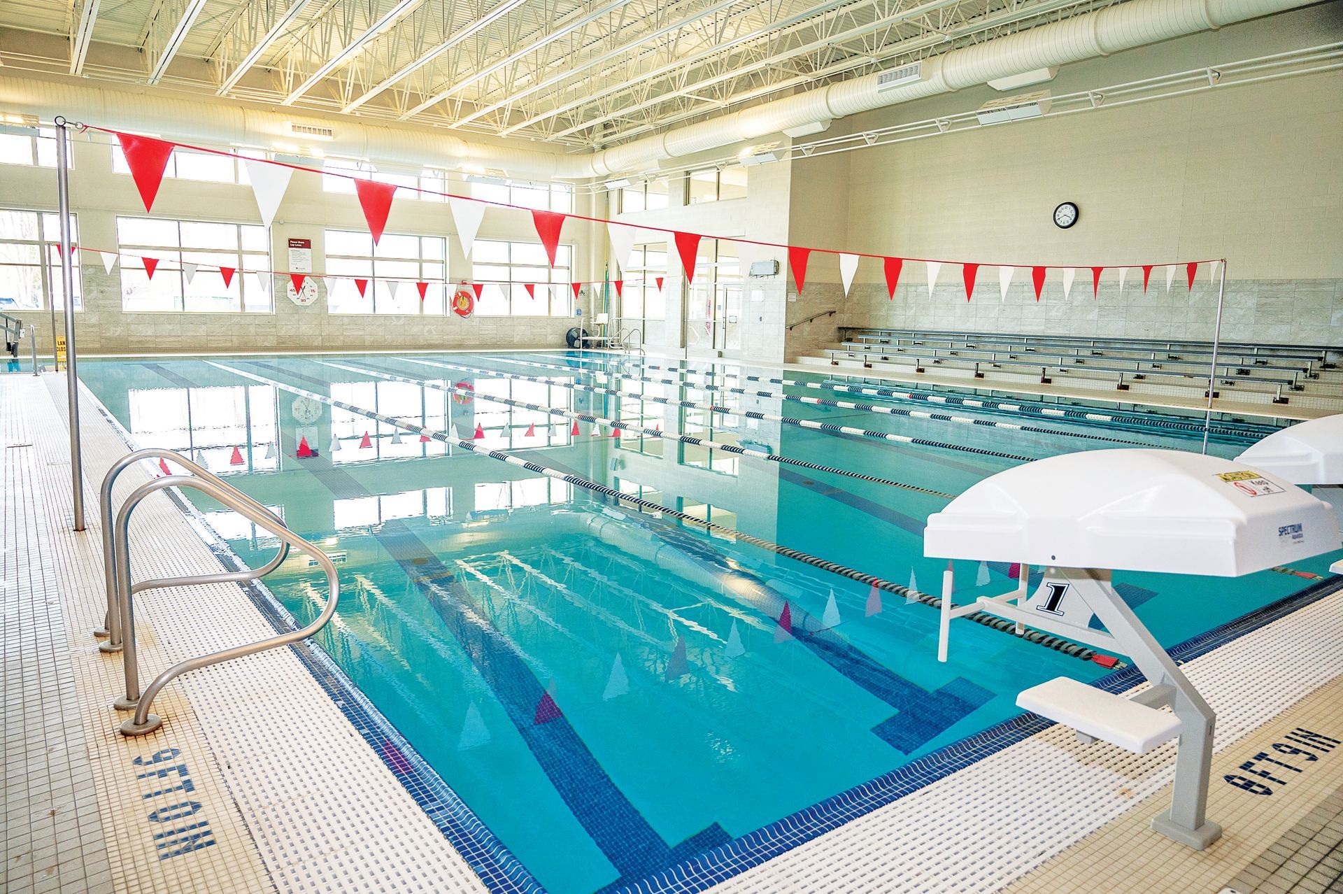 An indoor swimming pool with lap lanes, red and white flags overhead, starting blocks, and tile flooring.