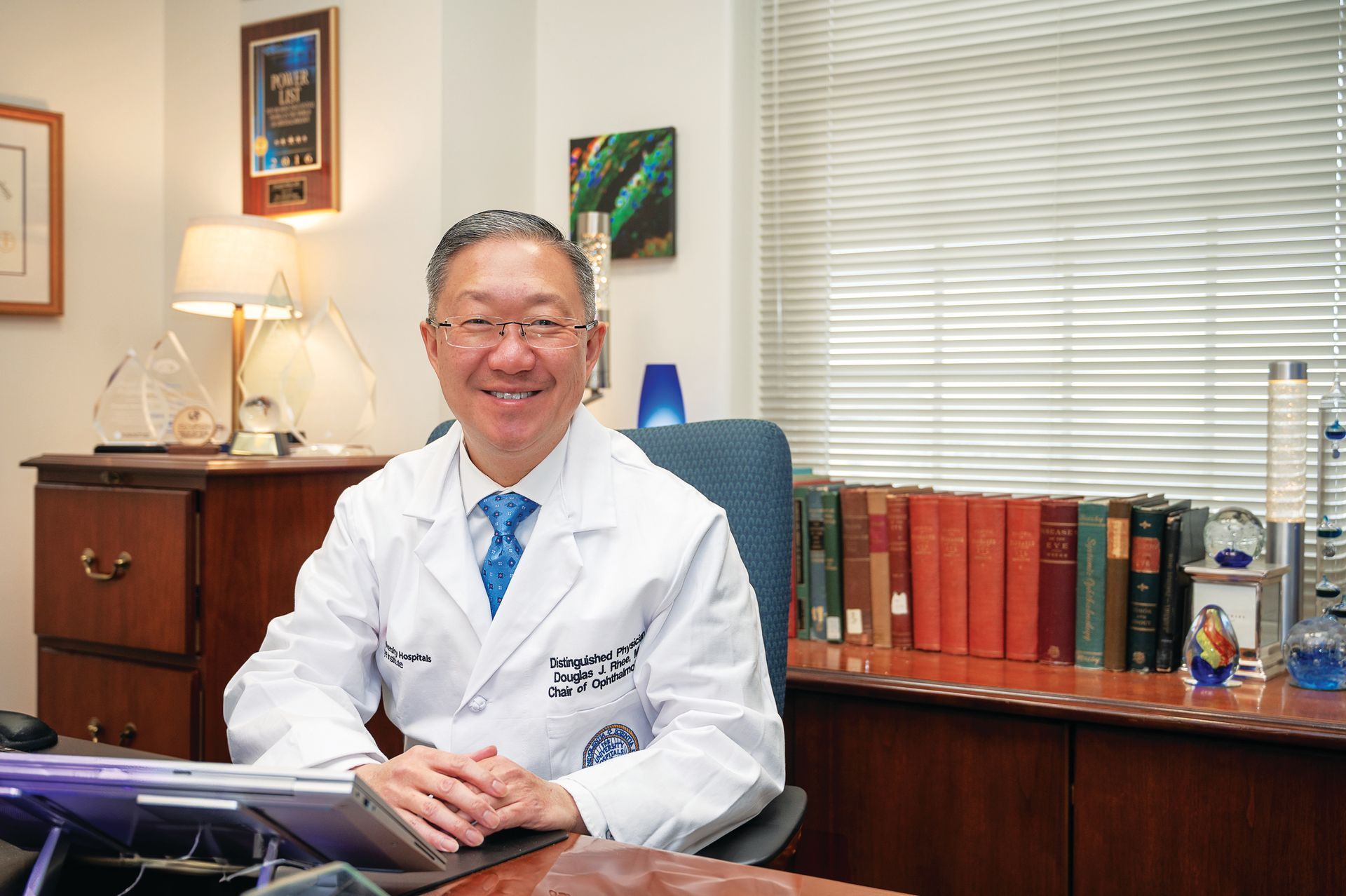 A smiling professional in a white lab coat sitting at a desk in an office with books and a table lamp.
