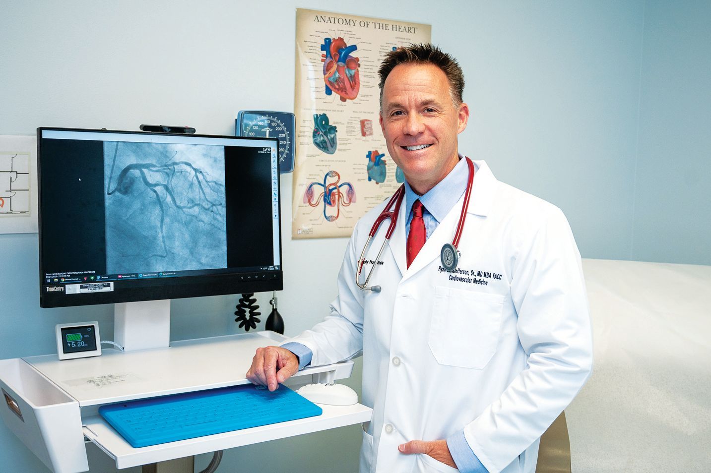 A doctor in a white coat stands next to a computer monitor showing a medical heart scan in an office with heart diagrams.
