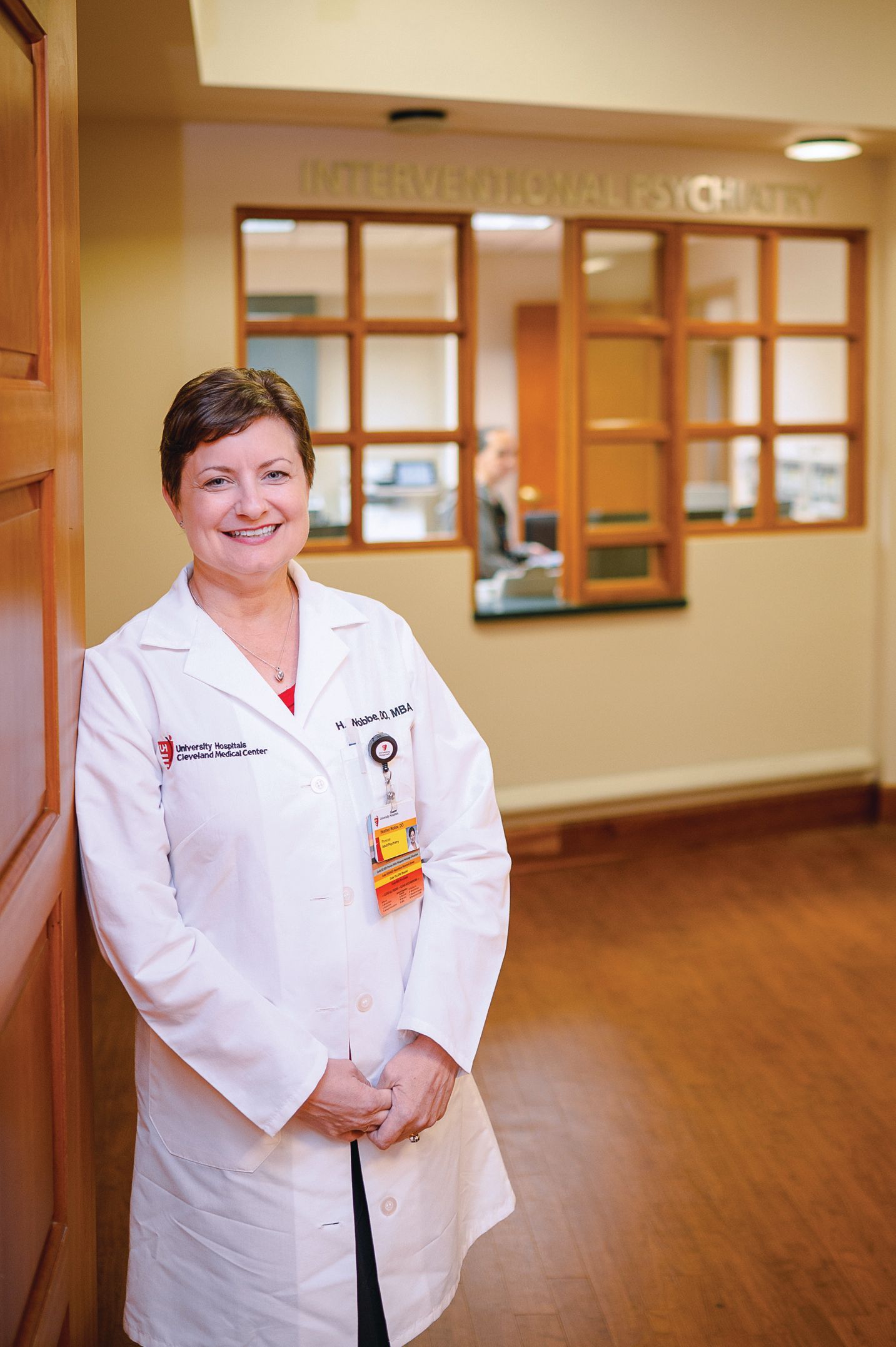 A healthcare professional in a white lab coat stands smiling in a brightly lit, wooden-toned office hallway.