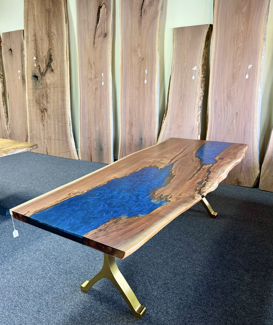 A live-edge wood dining table with a blue resin river inlay and gold metal Y-shaped legs, shown in a wood slab showroom.