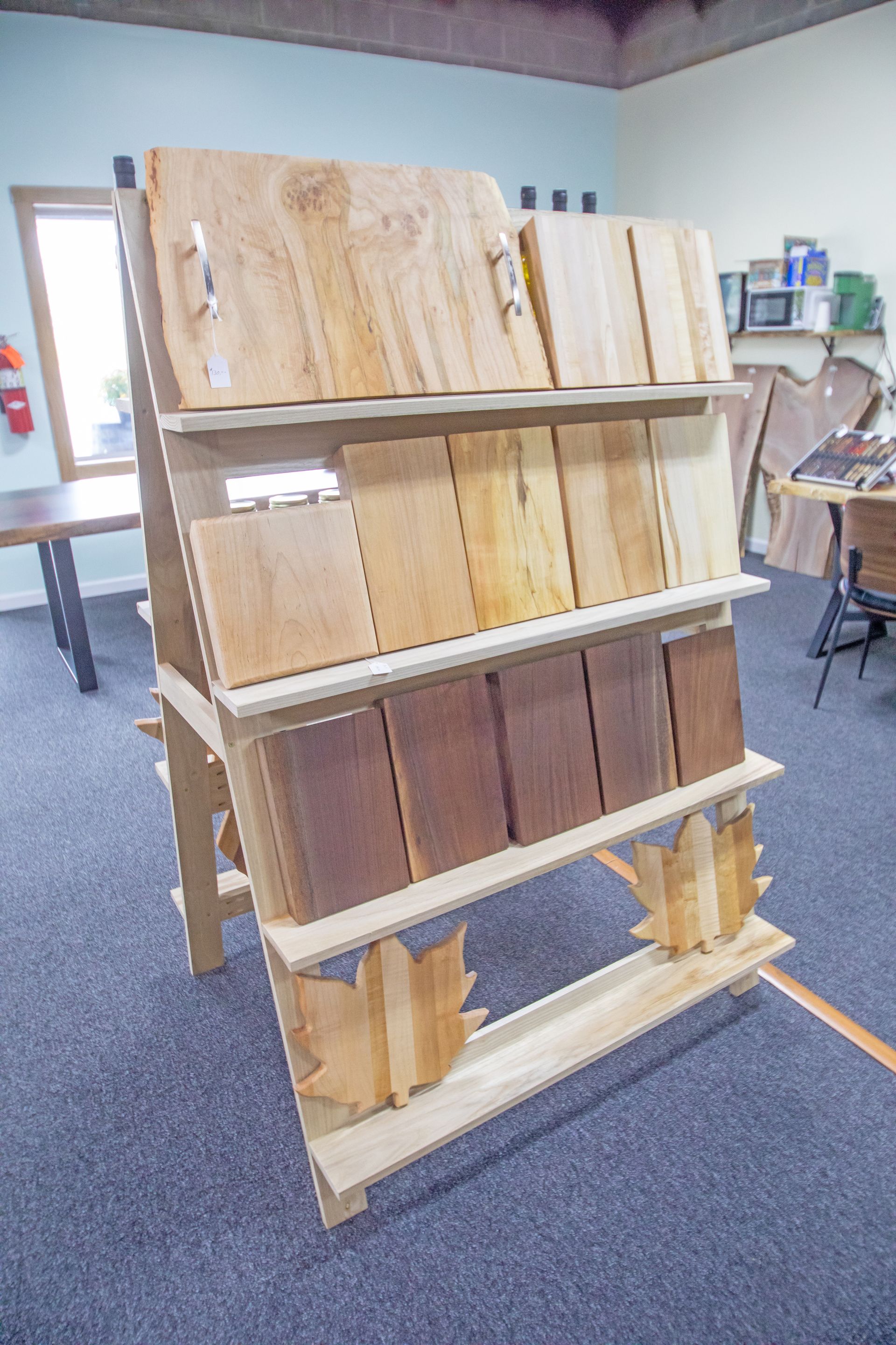 A wooden display rack holds various handcrafted wooden cutting boards and maple leaf-shaped decorations in a shop.