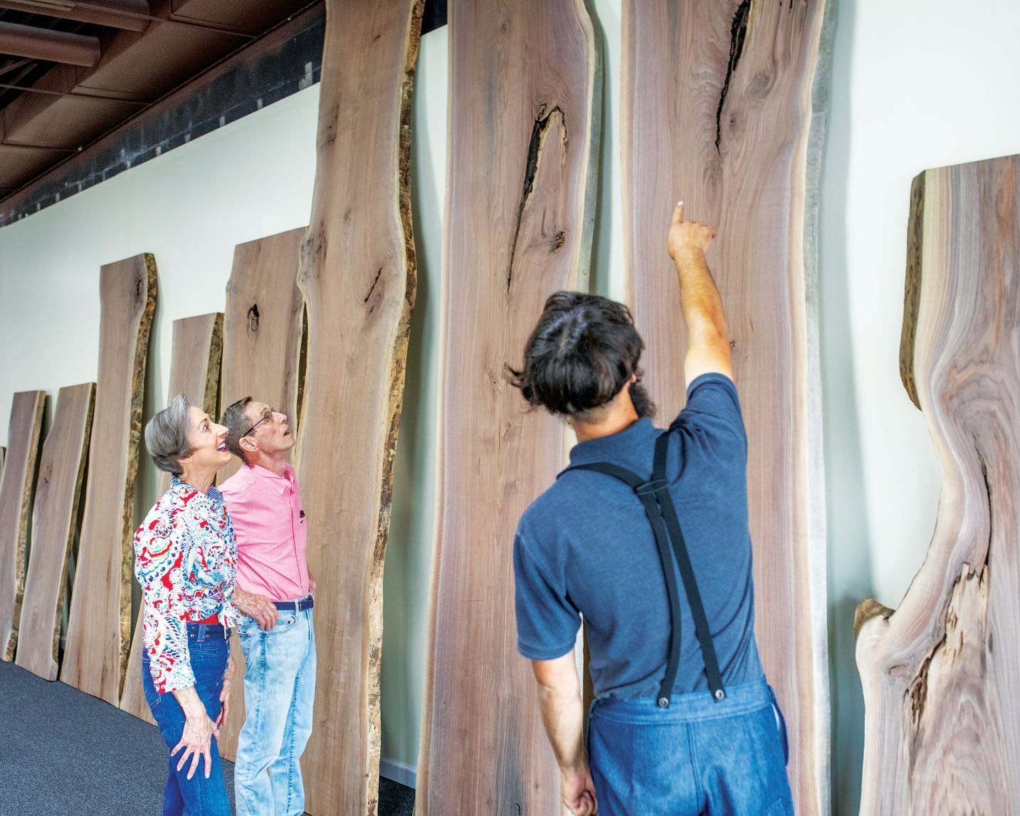 A person in overalls gestures toward large wooden slabs hanging on a wall, while two others look on in an indoor space.