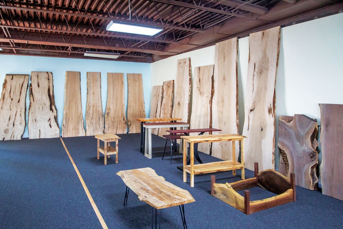 Large wooden slabs lean against walls in a showroom, with various handmade wooden tables and a pet bed on the floor.