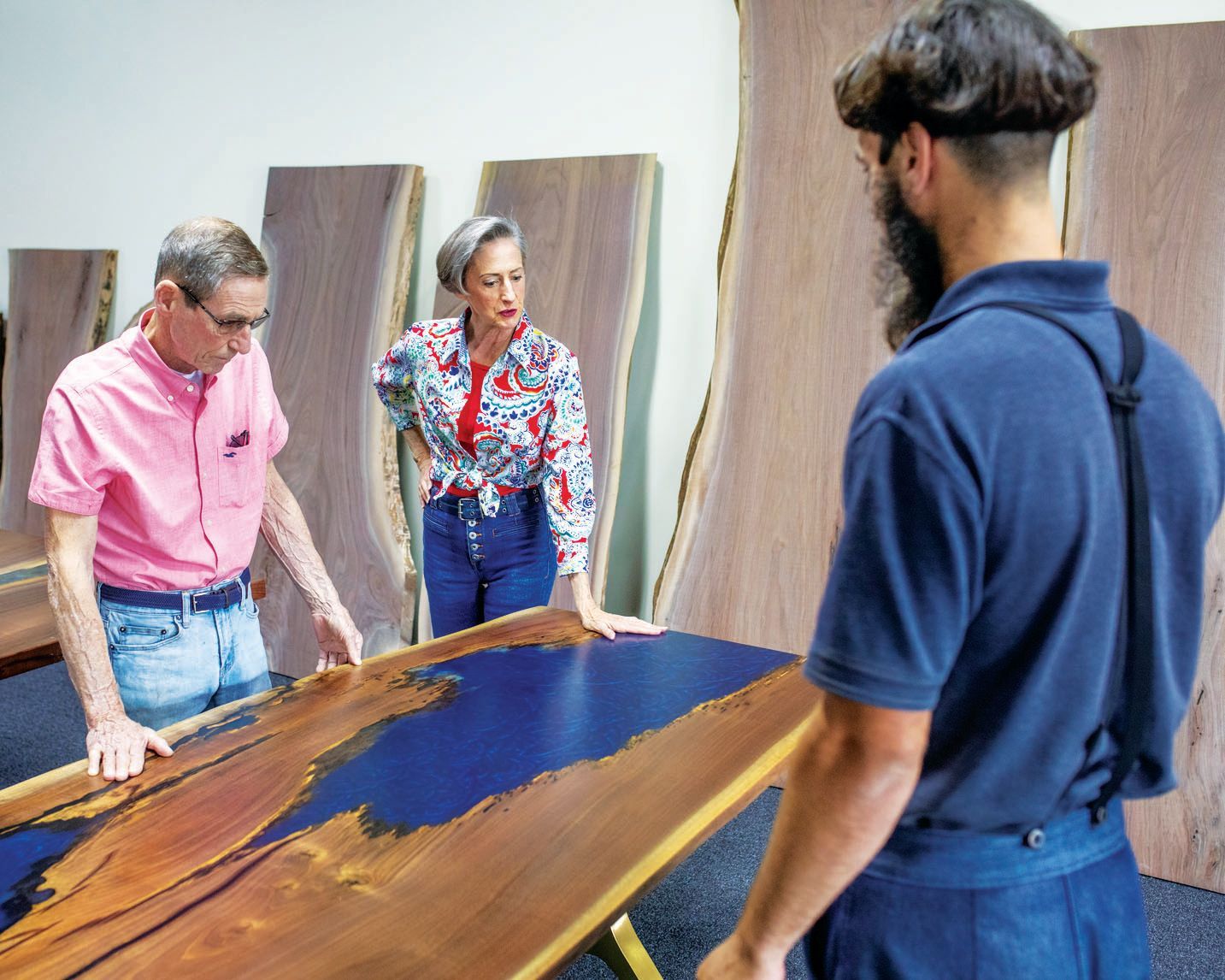Three people gather around a large live-edge wood table with a blue epoxy center in a workshop with wood slabs.