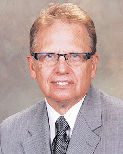 Professional headshot of a person wearing glasses, a gray suit, white collared shirt, and a dark patterned tie.