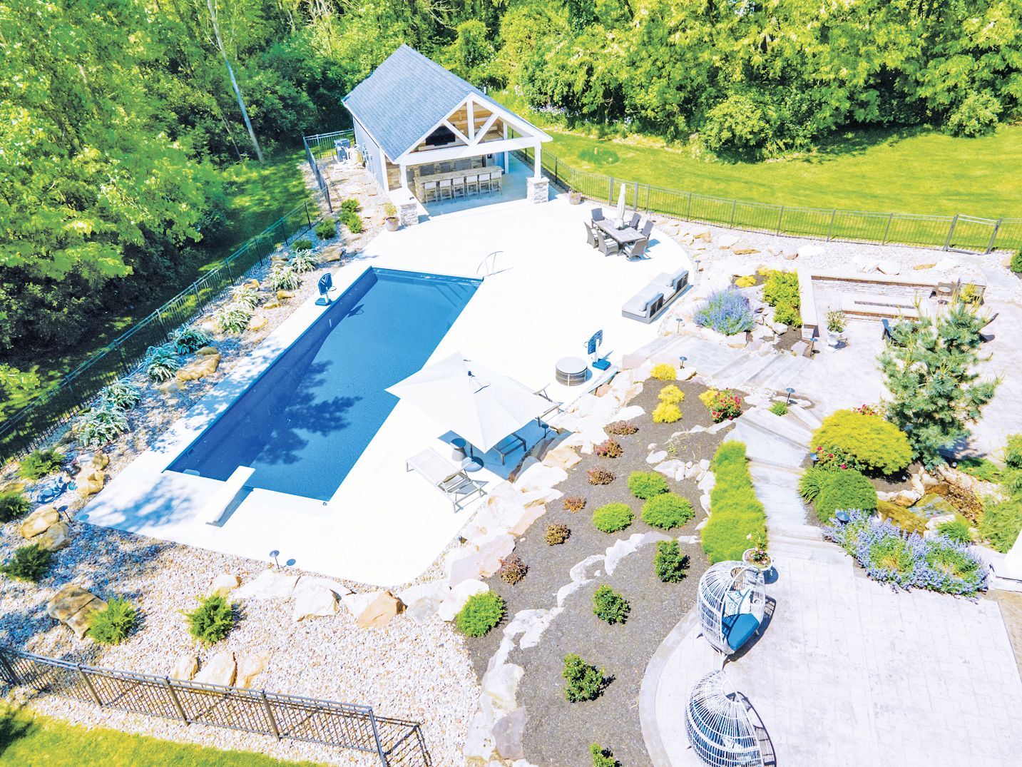 High-angle view of a backyard with an in-ground swimming pool, stone patio, lounge furniture, and a covered pavilion.