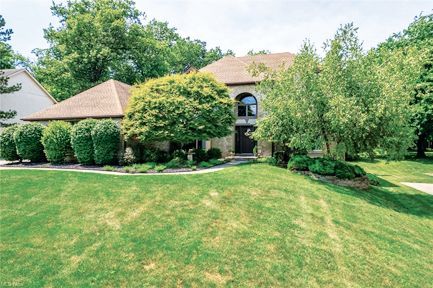 A two-story tan brick house with a brown roof, surrounded by lush green landscaping and trees under a clear blue sky.