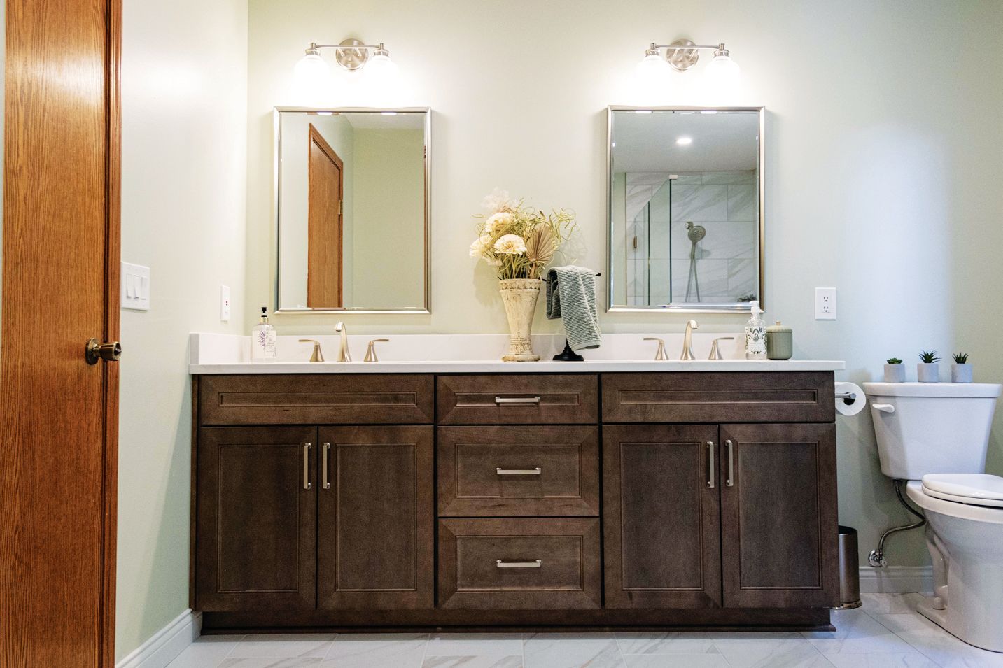 Double vanity in a bathroom with dark wood cabinets, white countertops, two sinks, mirrors, and a toilet to the right.