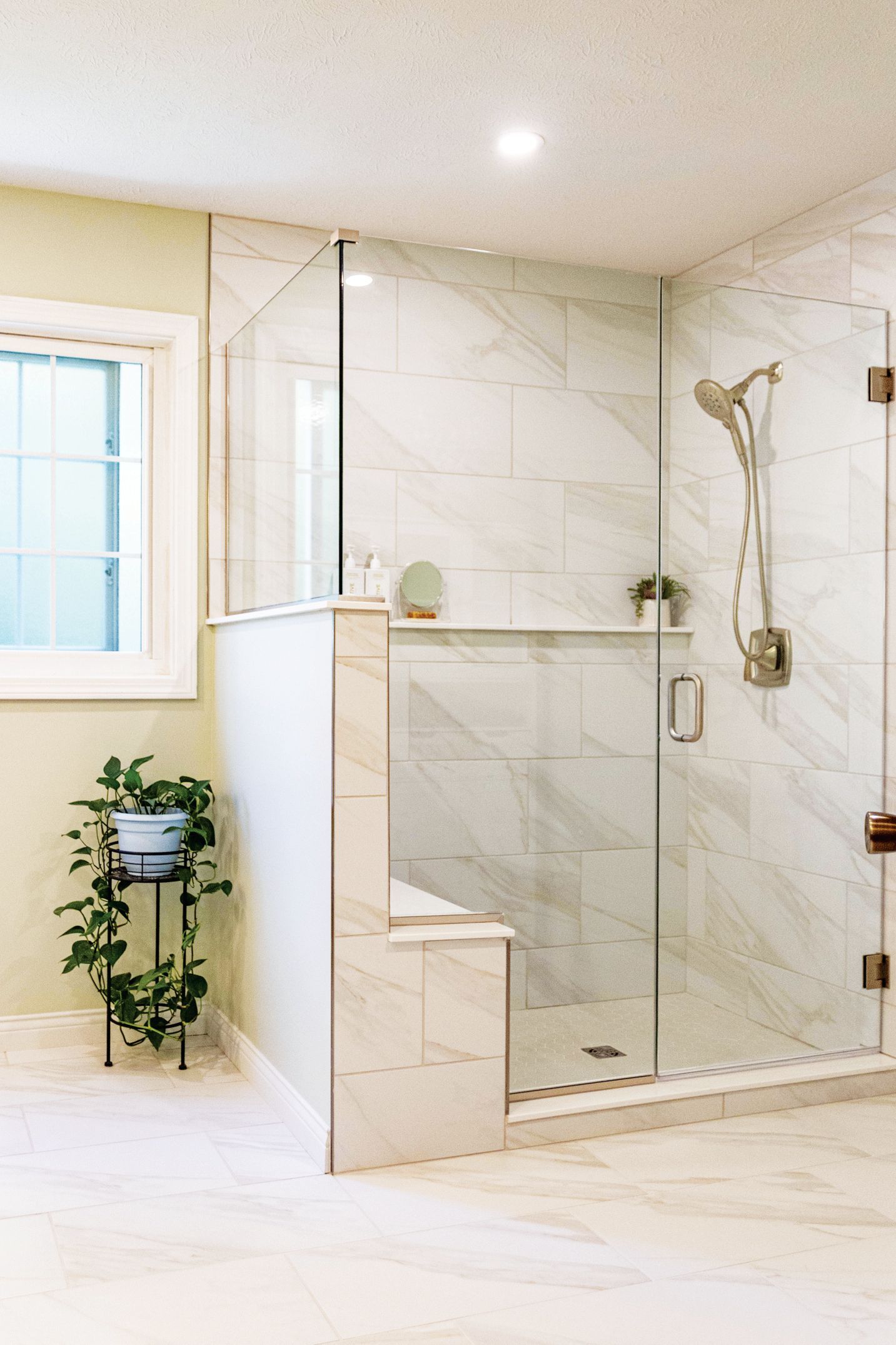 A modern walk-in shower with white marble tiles, glass doors, and a bench, next to a window and a potted plant.