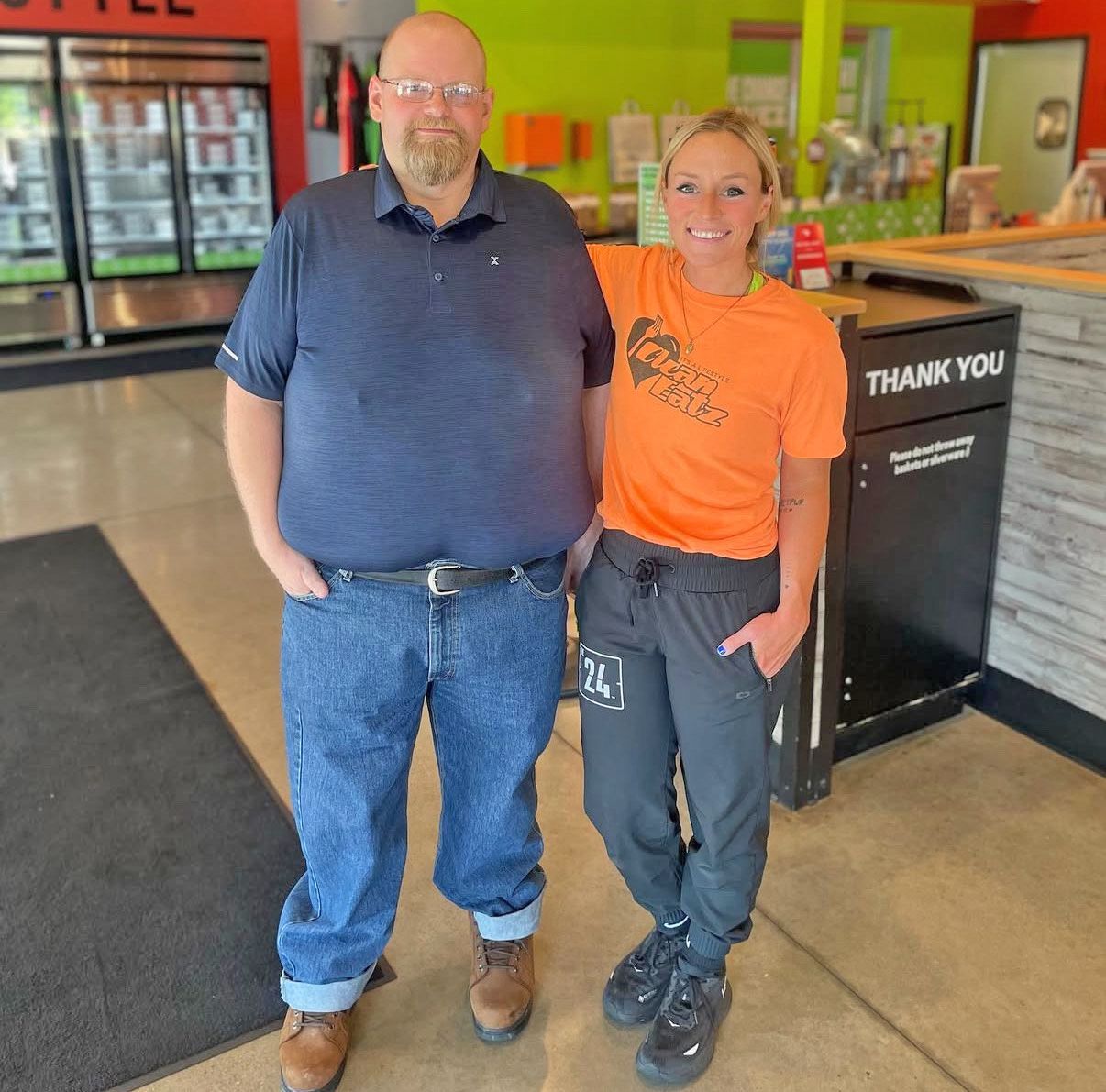 A man in a blue polo and jeans and a woman in an orange t-shirt and grey joggers pose for a photo in a cafe.