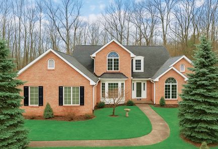 A two-story brick house with a dark shingled roof, arched windows, and a curved sidewalk in front of a forest.