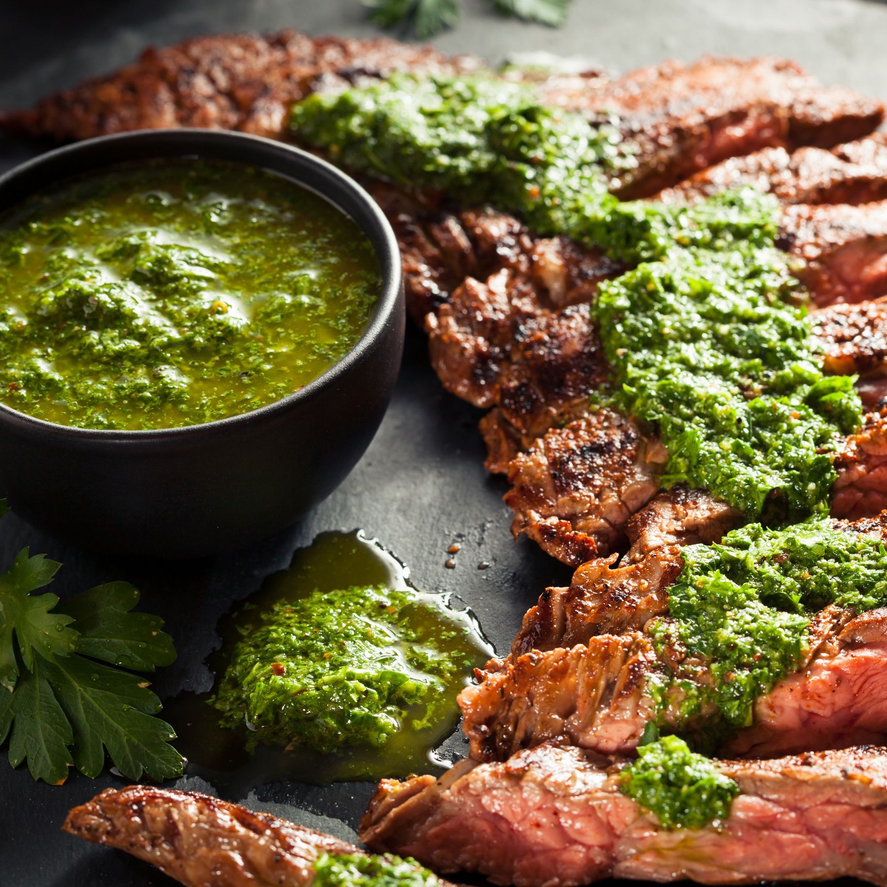 Slices of grilled steak topped with vibrant green chimichurri sauce, served with a small bowl of the sauce on a dark slate.