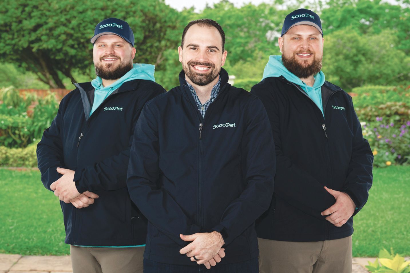 Three smiling professionals wearing branded dark jackets and matching caps stand outdoors in a grassy park setting.