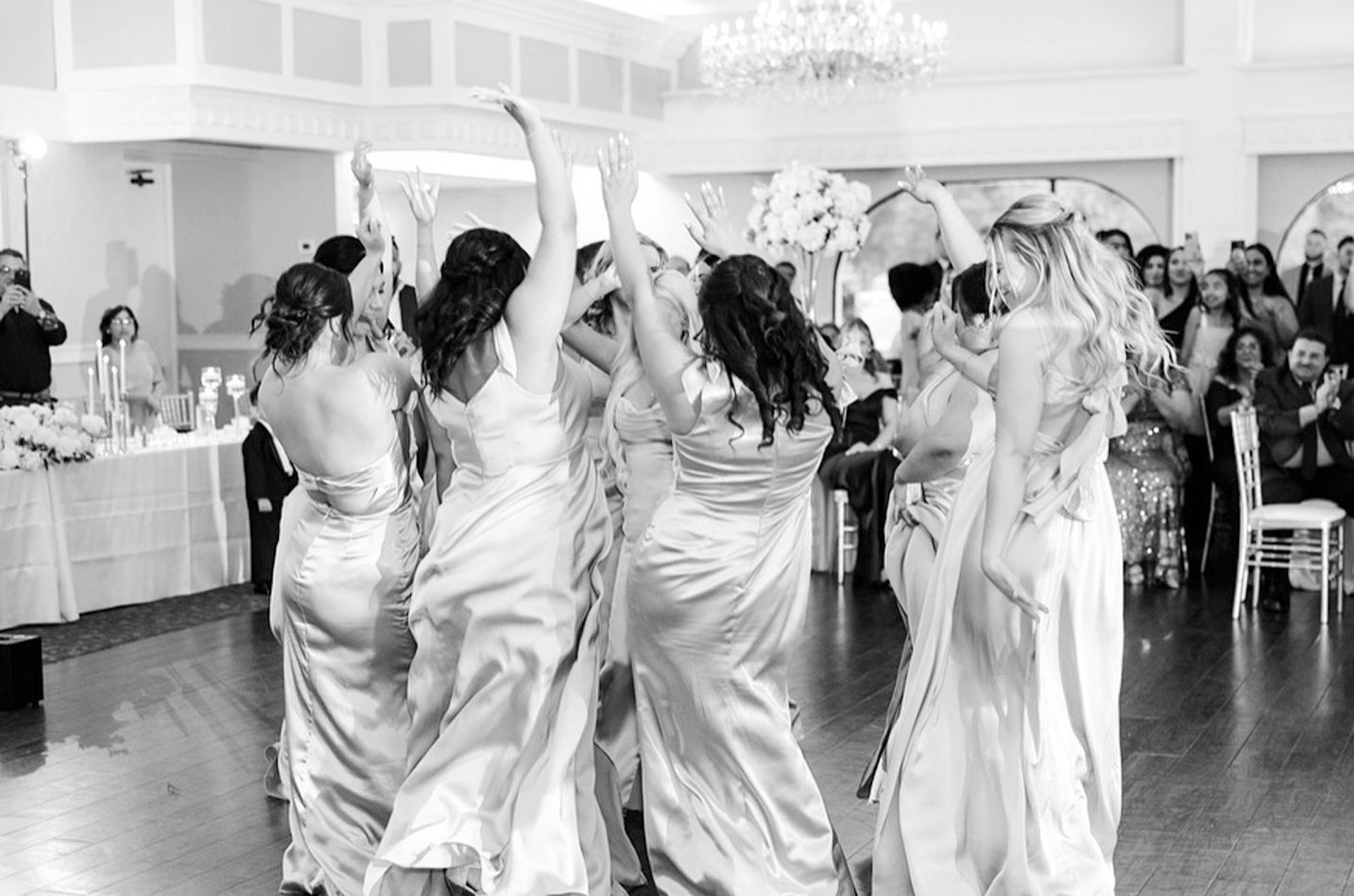 A group of people in formal gowns dancing in a reception hall with arms raised, celebrating at a wedding.