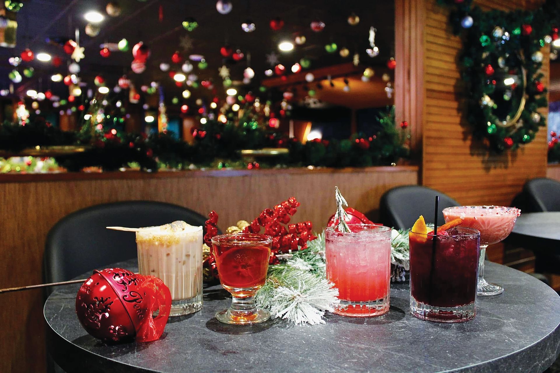 Five holiday-themed cocktails on a round table, surrounded by Christmas decorations against a festive bar background.