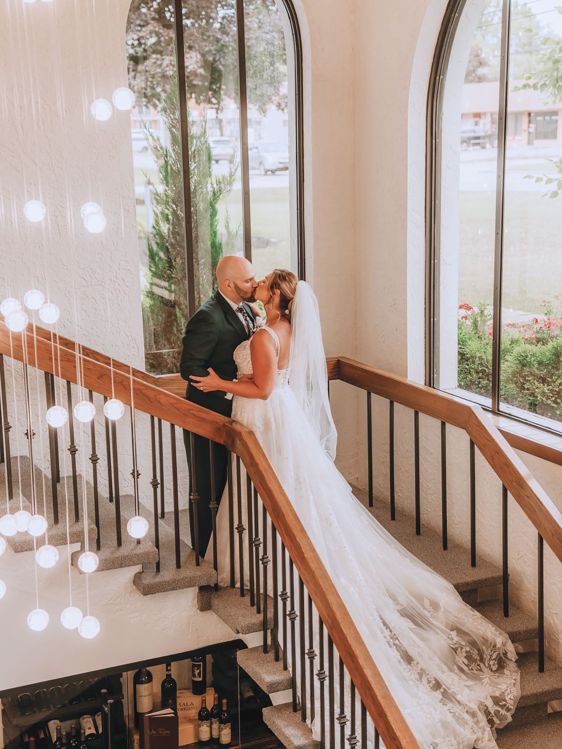 A couple in wedding attire kisses while standing on a staircase by a large arched window with a crystal chandelier.