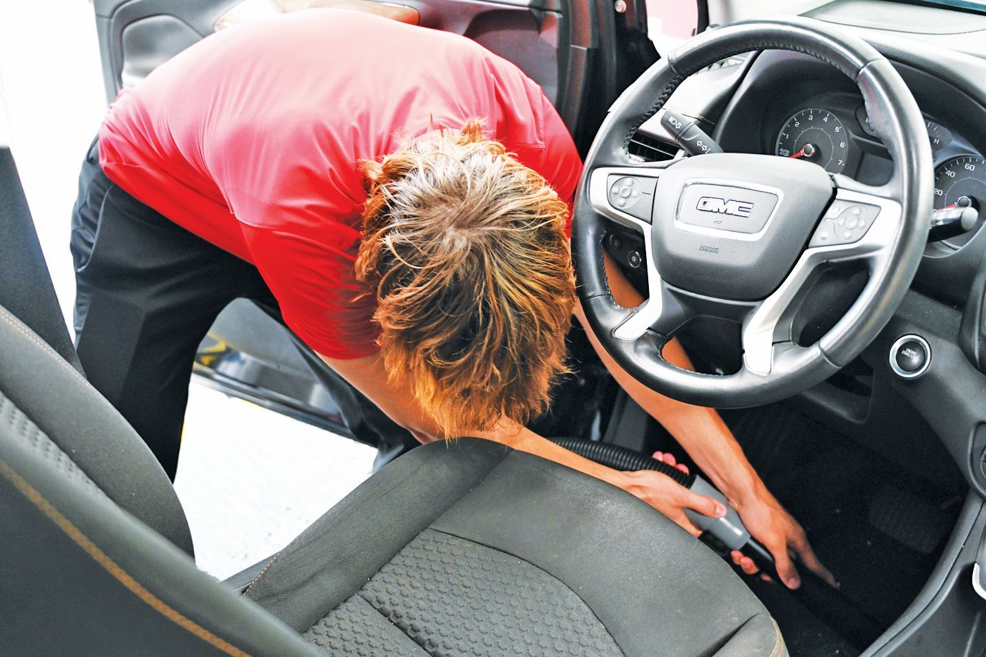 A person in a red shirt leans into the driver's side of a vehicle, vacuuming the floor near the pedals.