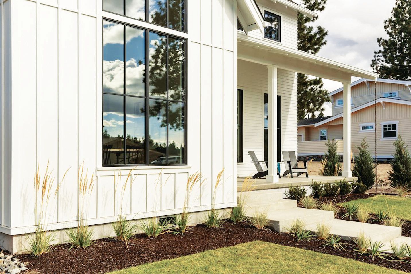 A modern white farmhouse with vertical siding, large black-framed windows, and a covered front porch with chairs.