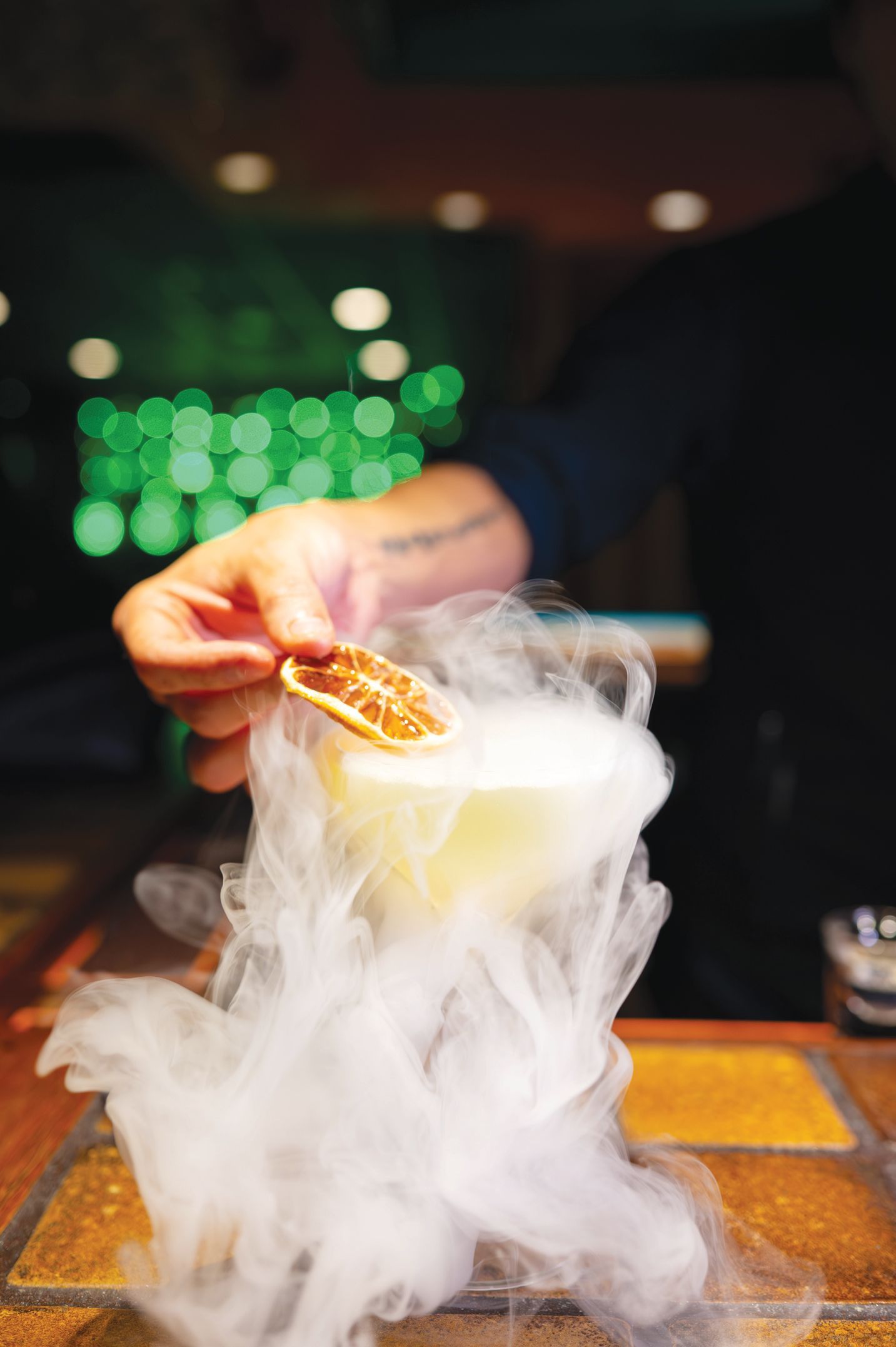 A person places a dried citrus slice on a cocktail glass emitting a thick, swirling fog of dry ice on a rustic counter.