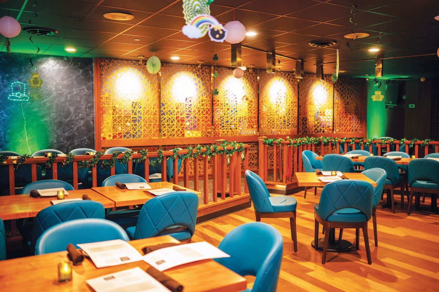 A restaurant interior with wooden tables, blue chairs, and walls decorated with gold mosaic panels and green lighting.