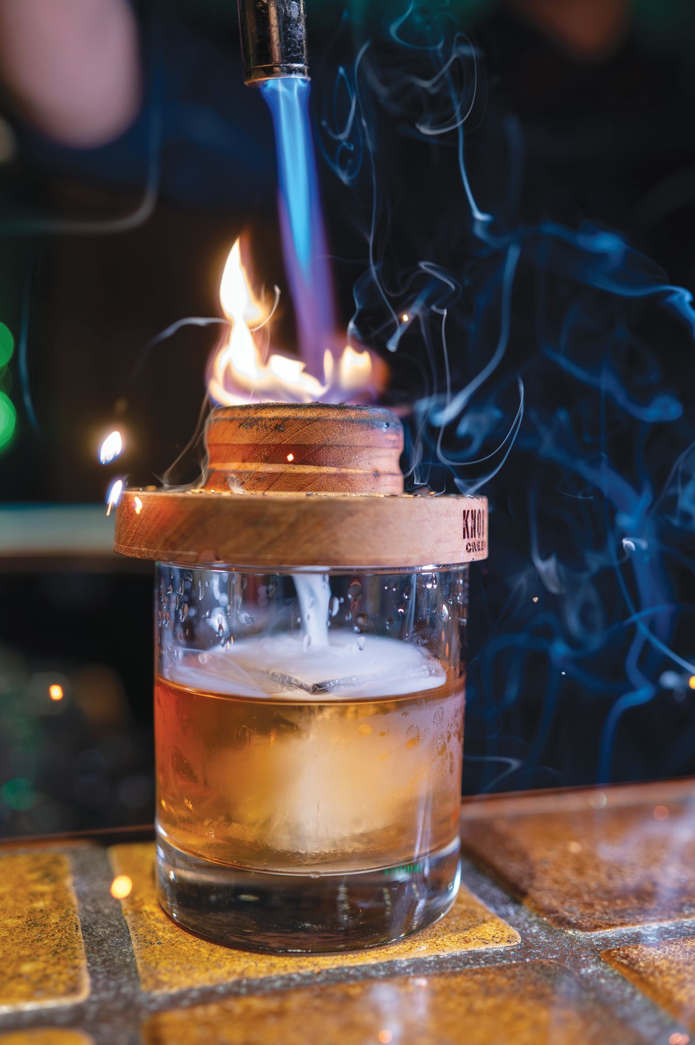 A cocktail being smoked with a kitchen torch on a wooden drink smoker board atop a glass filled with an amber liquid.