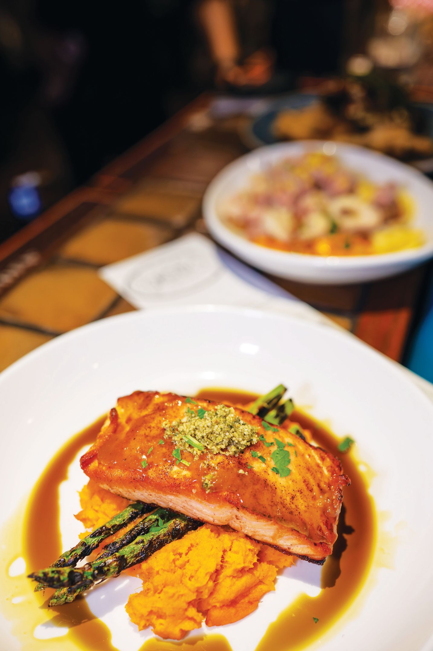 A salmon fillet served with mashed sweet potatoes and asparagus in a savory sauce, with a blurry dish in the background.