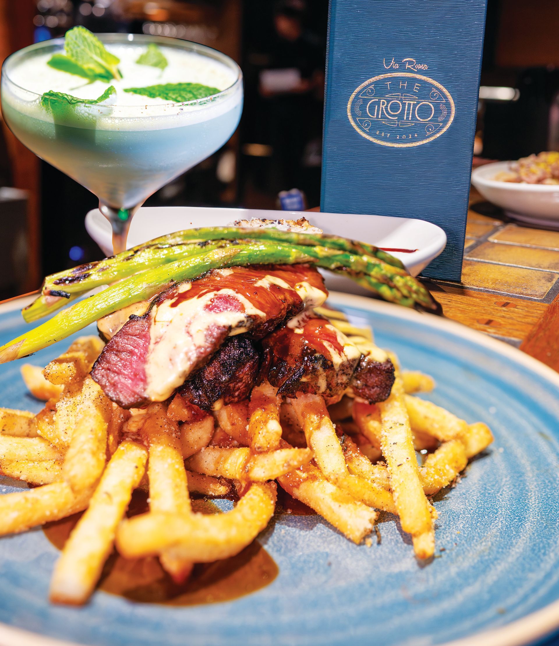 A blue plate topped with steak fries, grilled asparagus, and sauce, with a mint-garnished cocktail in the background.