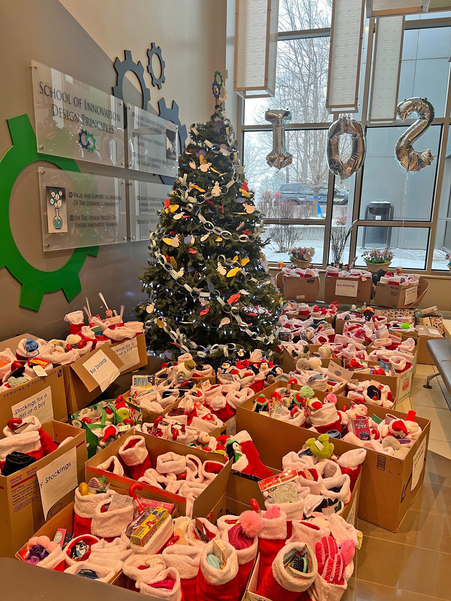 A Christmas tree surrounded by many cardboard boxes filled with red and white stockings in an indoor, lit office space.