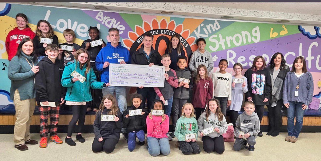 A group stands before a mural holding a large check, smiling in a school hallway.