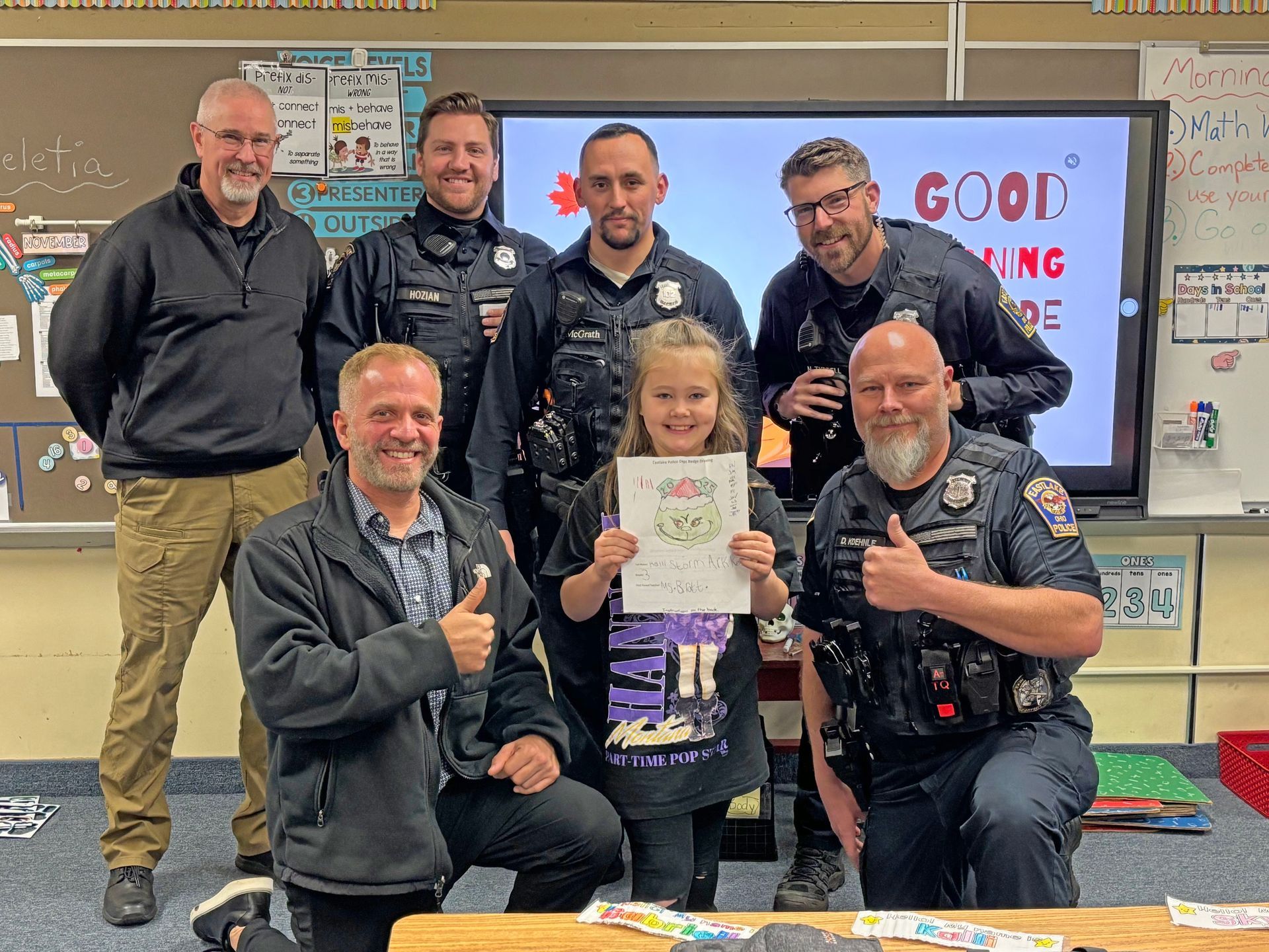 A group of uniformed officers and adults standing with a student holding a decorated paper in a classroom.
