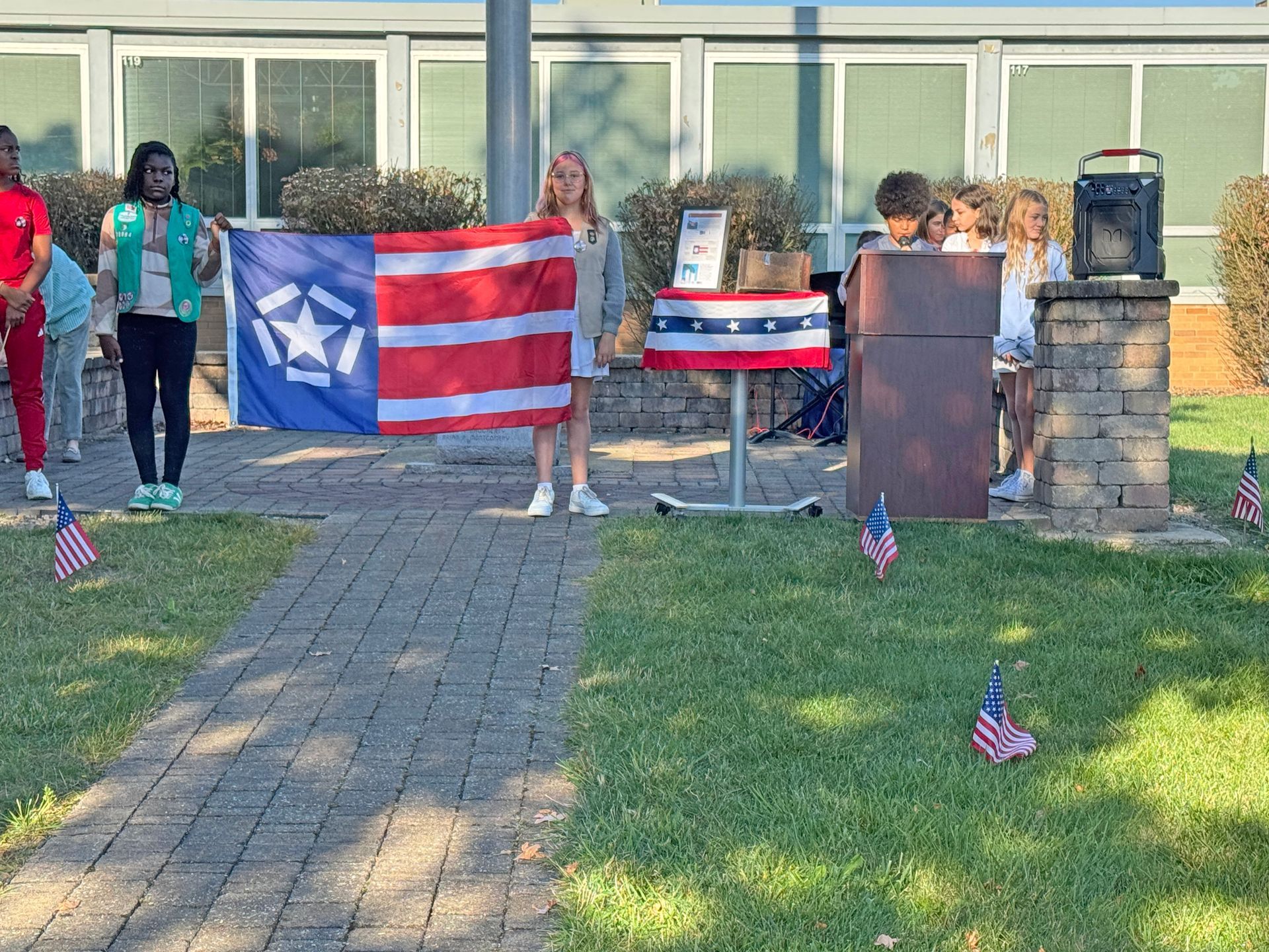 People holding a flag with a blue canton containing a star and stripes, standing outdoors near a podium and US flags.
