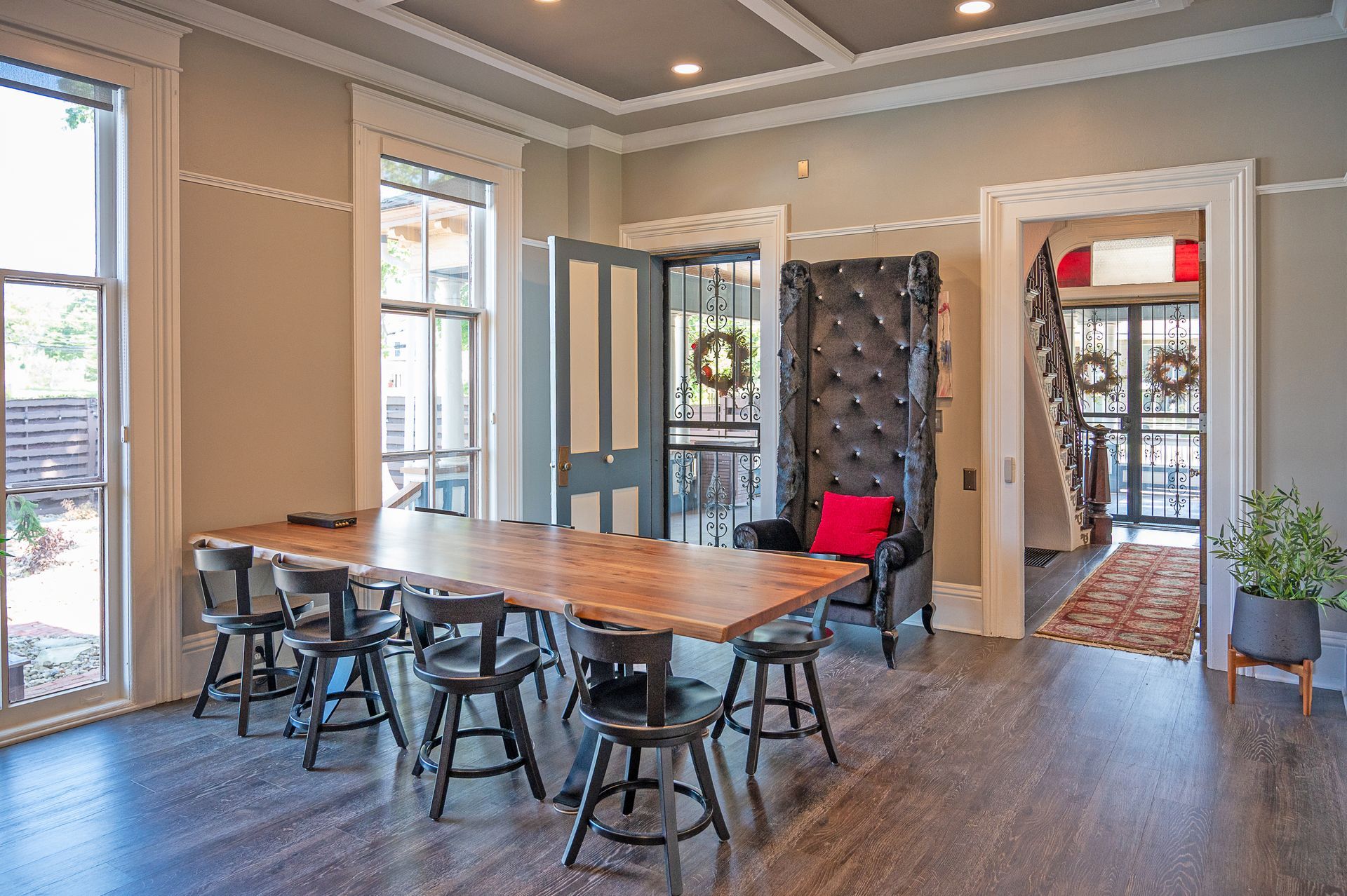 A long wooden dining table with stools in a room with a high-backed tufted chair, hardwood floors, and open doorways.