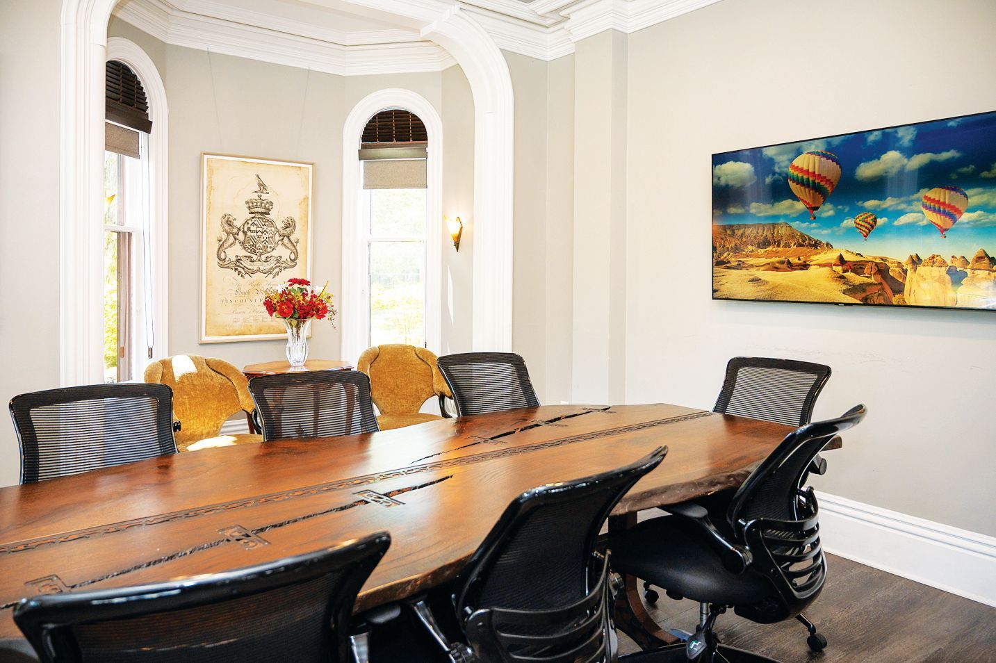 A wooden boardroom table with black office chairs, surrounded by light-colored walls and a wall-mounted hot air balloon art.