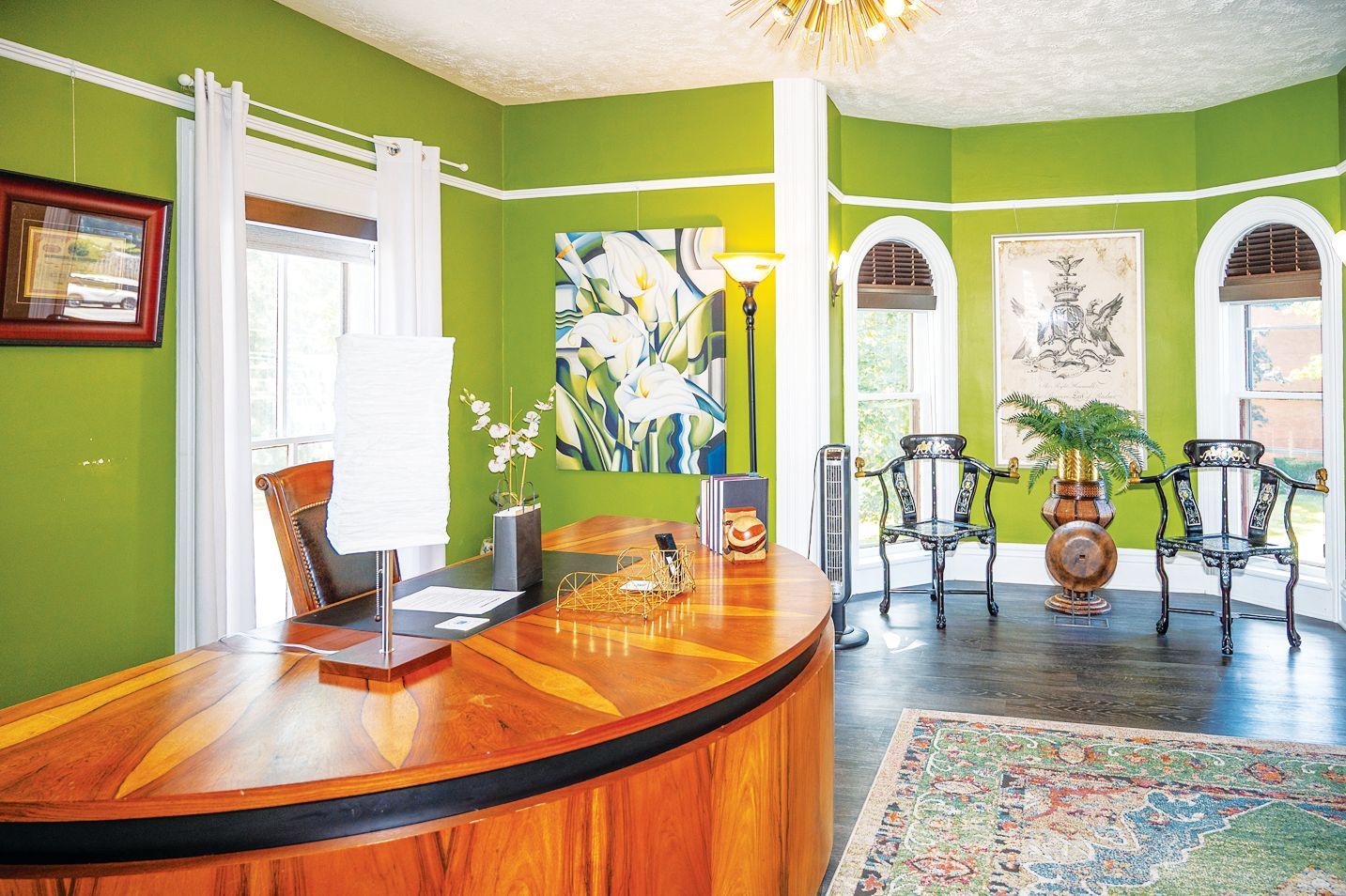 A bright lime-green office features a large, curved wooden desk, an ornate rug, and two chairs in a sunlit bay window area.