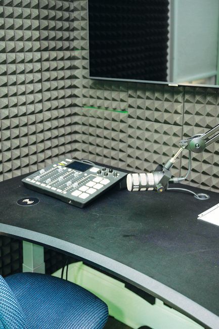 A radio studio setup with a sound mixing console and a microphone on a black desk against acoustic foam wall panels.