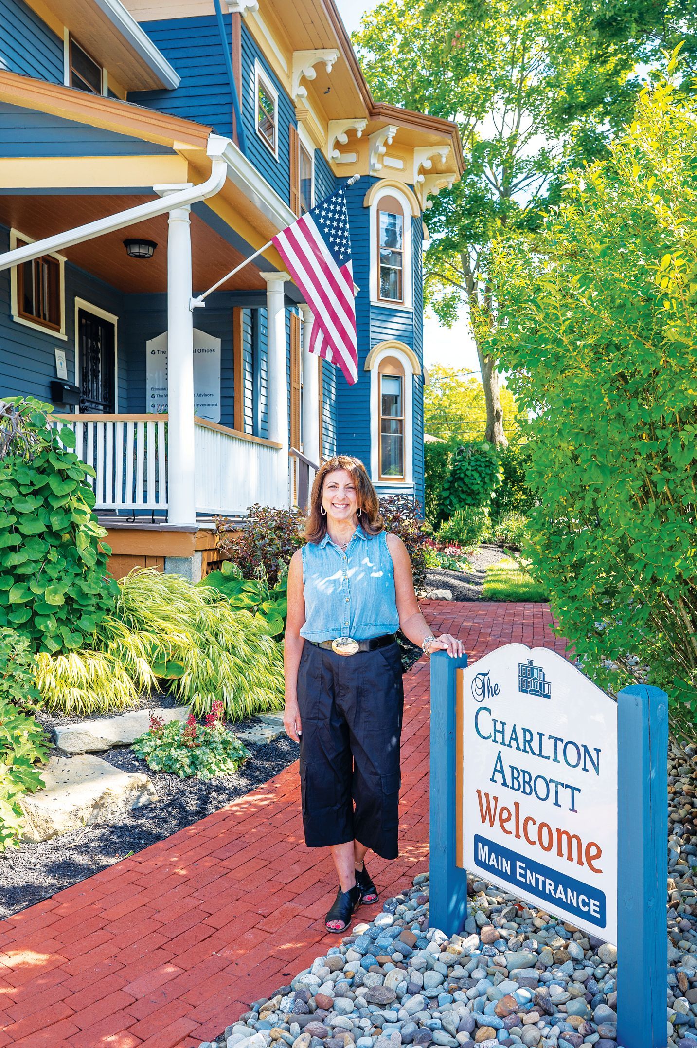A person stands smiling on a brick walkway in front of a blue Victorian house with an American flag and a welcome sign.
